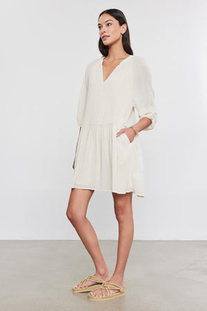 A woman in a white Velvet by Graham & Spencer Sloan Dress with pleat detailing and beige sandals stands against a gray background, looking to the side with her hands slightly behind her.