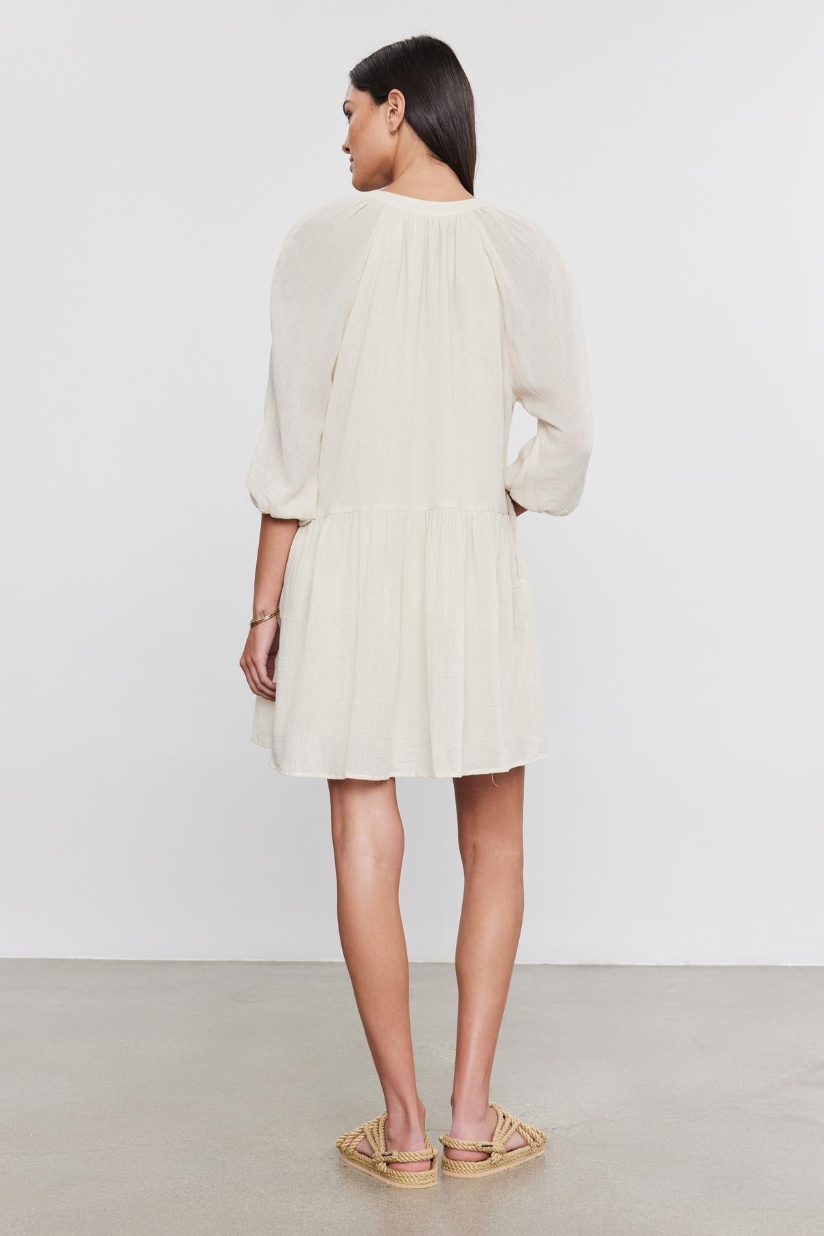 A woman standing and facing away from the camera, wearing a short, pale beige Velvet by Graham & Spencer Sloan Dress with pleat detailing, puffed sleeves, and open-toe sandals.-36752957112513