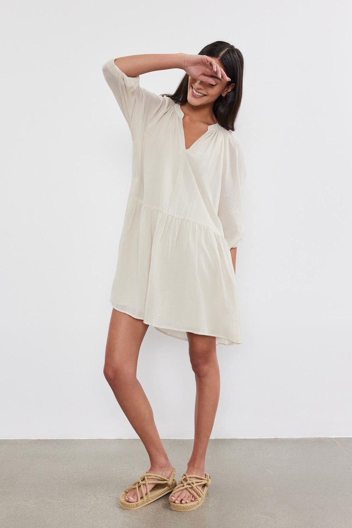   Woman in a beige Velvet by Graham & Spencer Sloan Dress with pleat detailing, shielding her eyes with her hand, standing against a white background. 