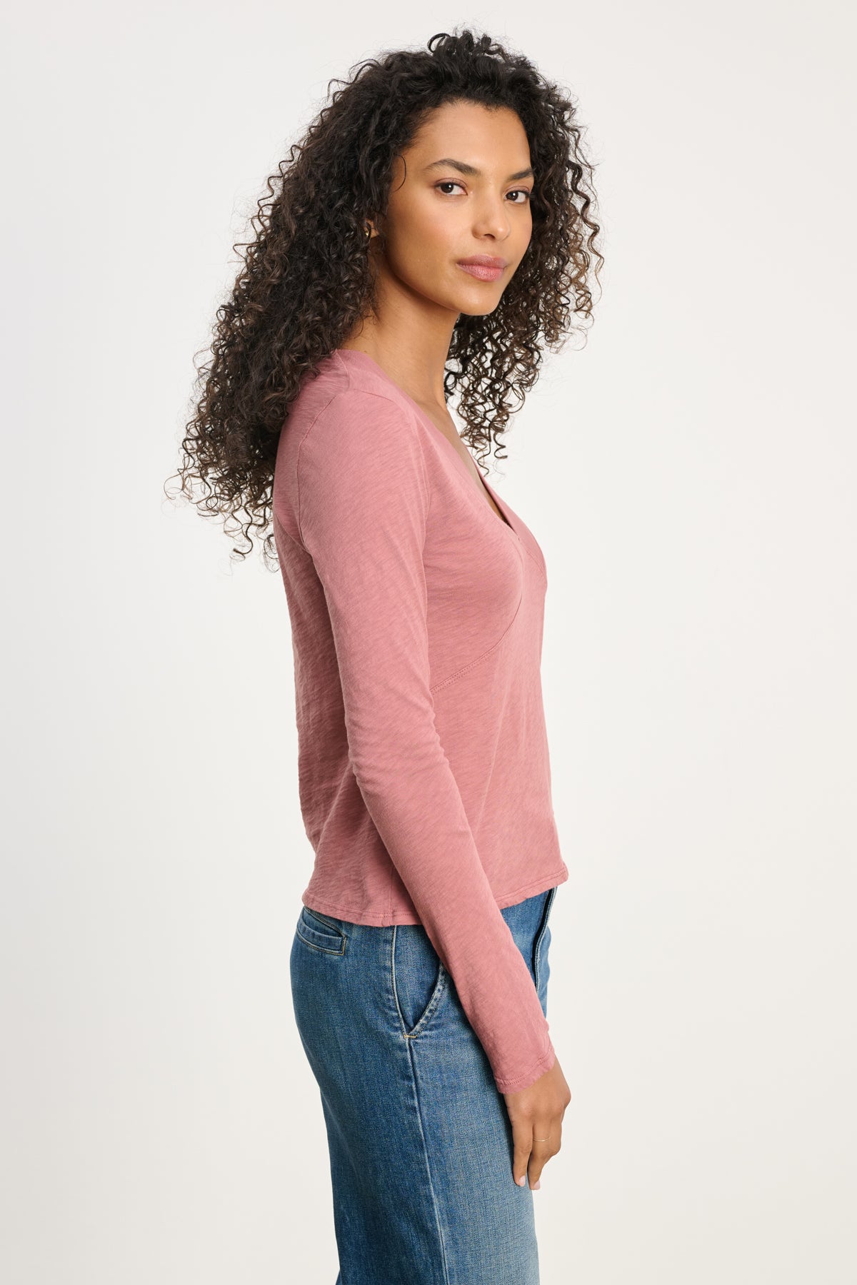   A woman with curly hair stands in profile against a light background, wearing the Velvet by Graham & Spencer NESSA TEE in pink cotton slub with blue jeans, showcasing a relaxed modern look. 
