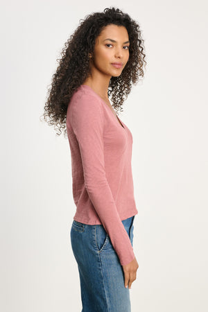 A woman with curly hair stands in profile against a light background, wearing the Velvet by Graham & Spencer NESSA TEE in pink cotton slub with blue jeans, showcasing a relaxed modern look.