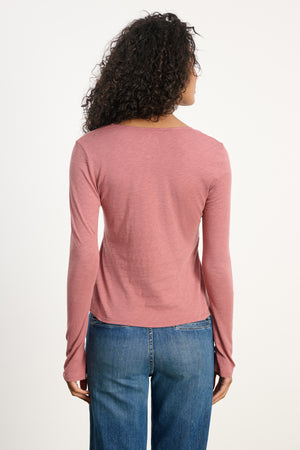 Person with curly hair, shown from the back in Velvet by Graham & Spencer's NESSA TEE—a vintage-inspired pink cotton slub long-sleeve top—paired with blue jeans against a plain light background for a relaxed modern vibe.