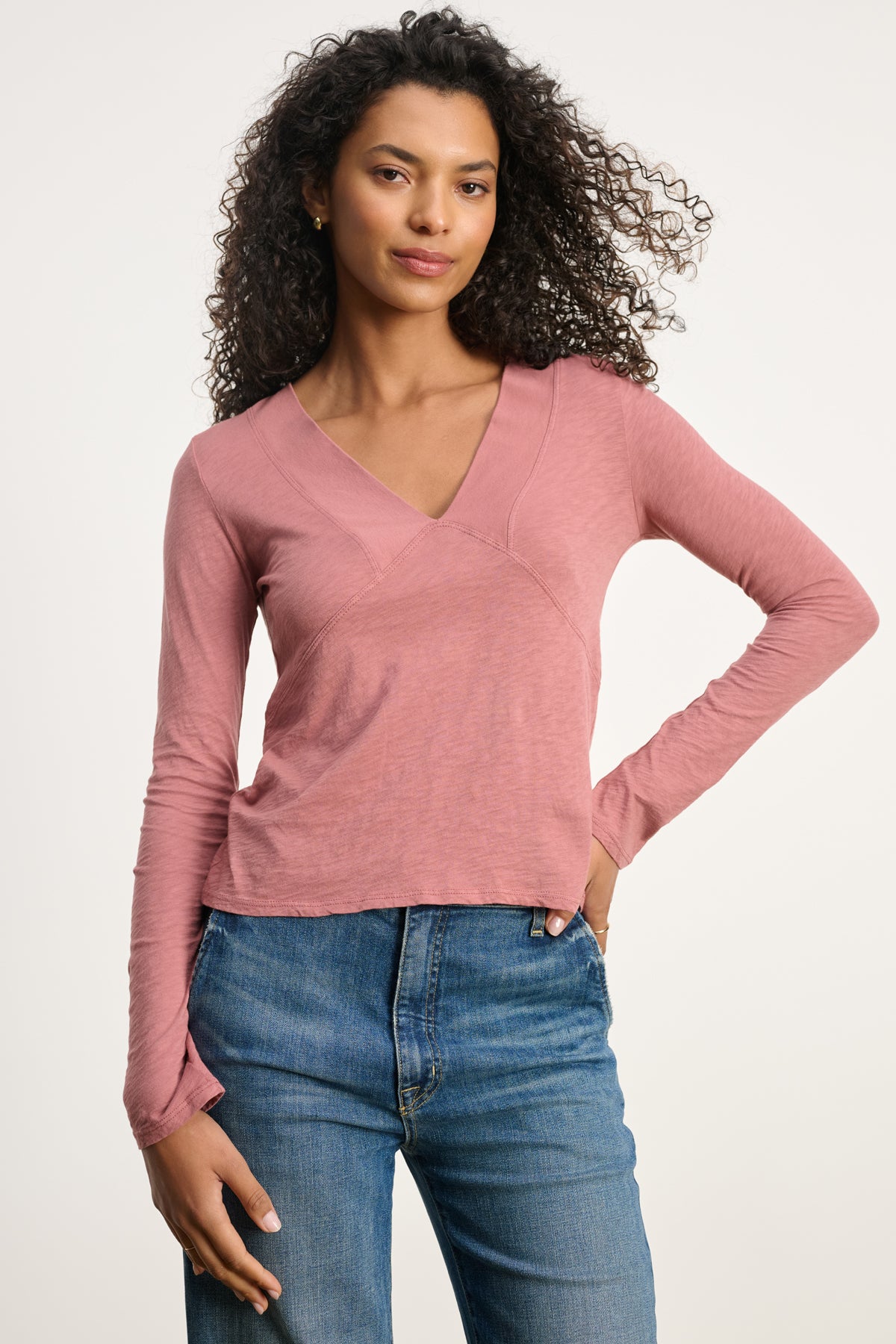 A woman with curly hair wears the NESSA TEE by Velvet by Graham & Spencer, a vintage cotton slub pink deep V-neck tee, paired with blue jeans, standing against a plain light background.-39838367023297