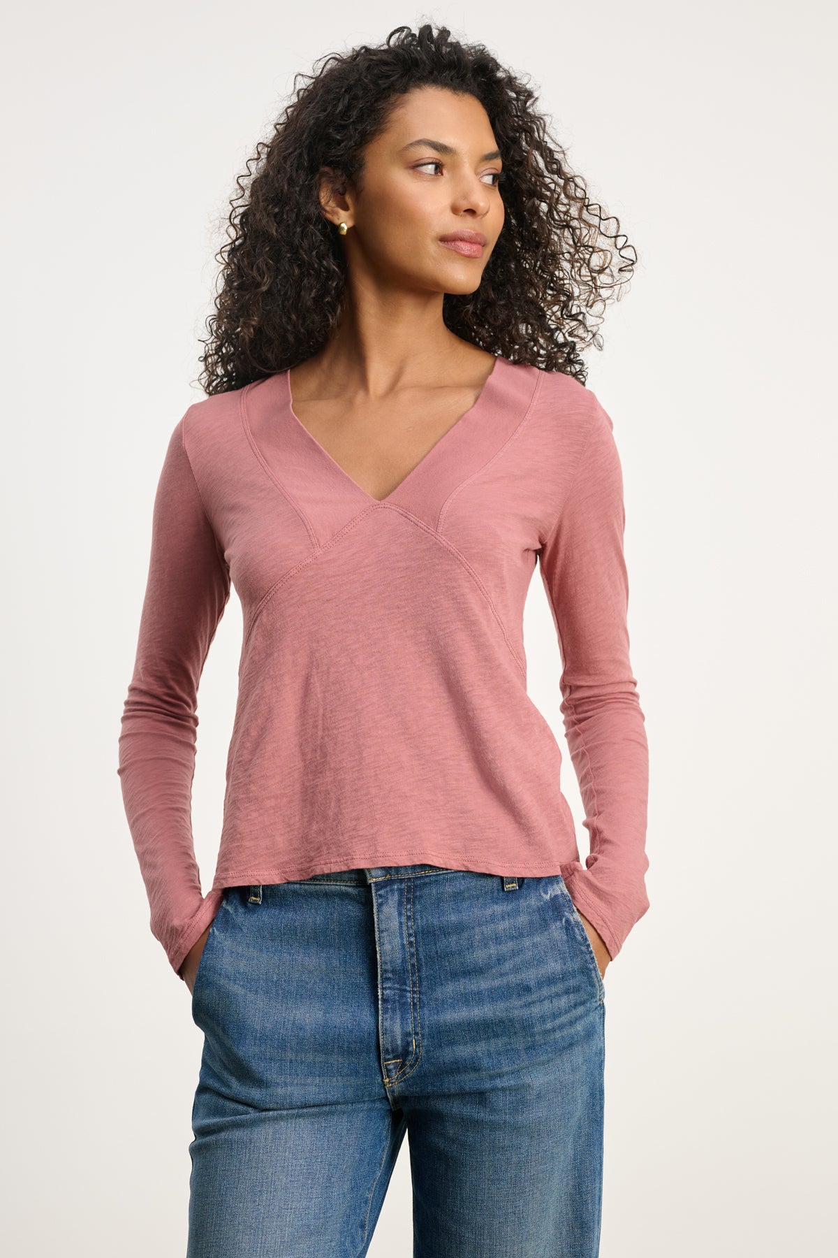   A woman wears the NESSA TEE by Velvet by Graham & Spencer with blue jeans, hands in her pockets, gazing to the side for a relaxed modern look against a plain background. 