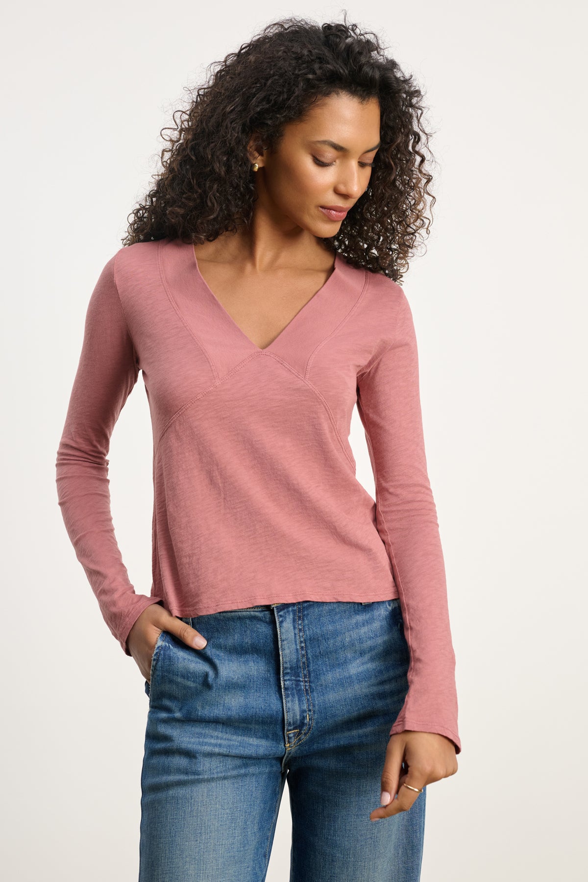 A woman wears the NESSA TEE by Velvet by Graham & Spencer, a vintage cotton slub deep V-neck in pink, with blue jeans against a plain light background for a relaxed modern look.-39838366957761