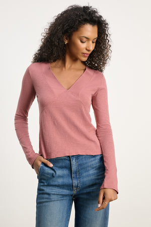 A woman wears the NESSA TEE by Velvet by Graham & Spencer, a vintage cotton slub deep V-neck in pink, with blue jeans against a plain light background for a relaxed modern look.