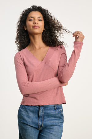 A woman with curly hair, dressed in the Velvet by Graham & Spencer NESSA TEE—a long-sleeve deep V-neck—paired with blue jeans, stands against a plain background, holding a strand of her hair.