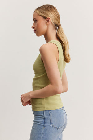 Side profile of a woman wearing a NOLA tank top by Velvet by Graham & Spencer in green and blue jeans, with her blond hair in a low ponytail, standing against a plain white background.