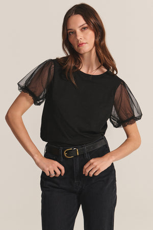 A woman wears the JUSTINE TEE by Velvet by Graham & Spencer—black cotton slub with sheer puff sleeves and a crew neck—paired with black pants, standing against a neutral background with one hand on her hip.