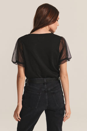 A woman stands with her back to the camera, wearing the Velvet by Graham & Spencer JUSTINE TEE— a black cotton slub top with sheer puff sleeves—paired with high-waisted black jeans, set against a neutral background.
