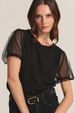 A woman wears the JUSTINE TEE by Velvet by Graham & Spencer— a black crew neck top with sheer mesh sleeves—paired with dark jeans and a black belt, standing against a neutral background.