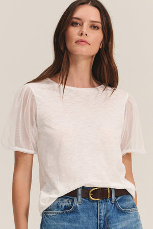 A woman wears the JUSTINE TEE by Velvet by Graham & Spencer—a white cotton slub top with sheer tulle puff sleeves and a classic crew neckline—paired with blue jeans and a dark belt, standing against a plain light background.