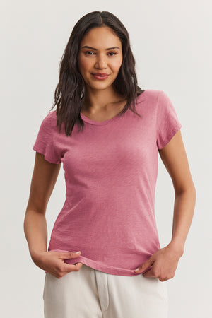 A person wears the Velvet by Graham & Spencer ODELIA TEE in pink, made from premium cotton slub, paired with light pants against a neutral background. The classic crew neckline enhances this wardrobe essential's timeless appeal as they look at the camera.