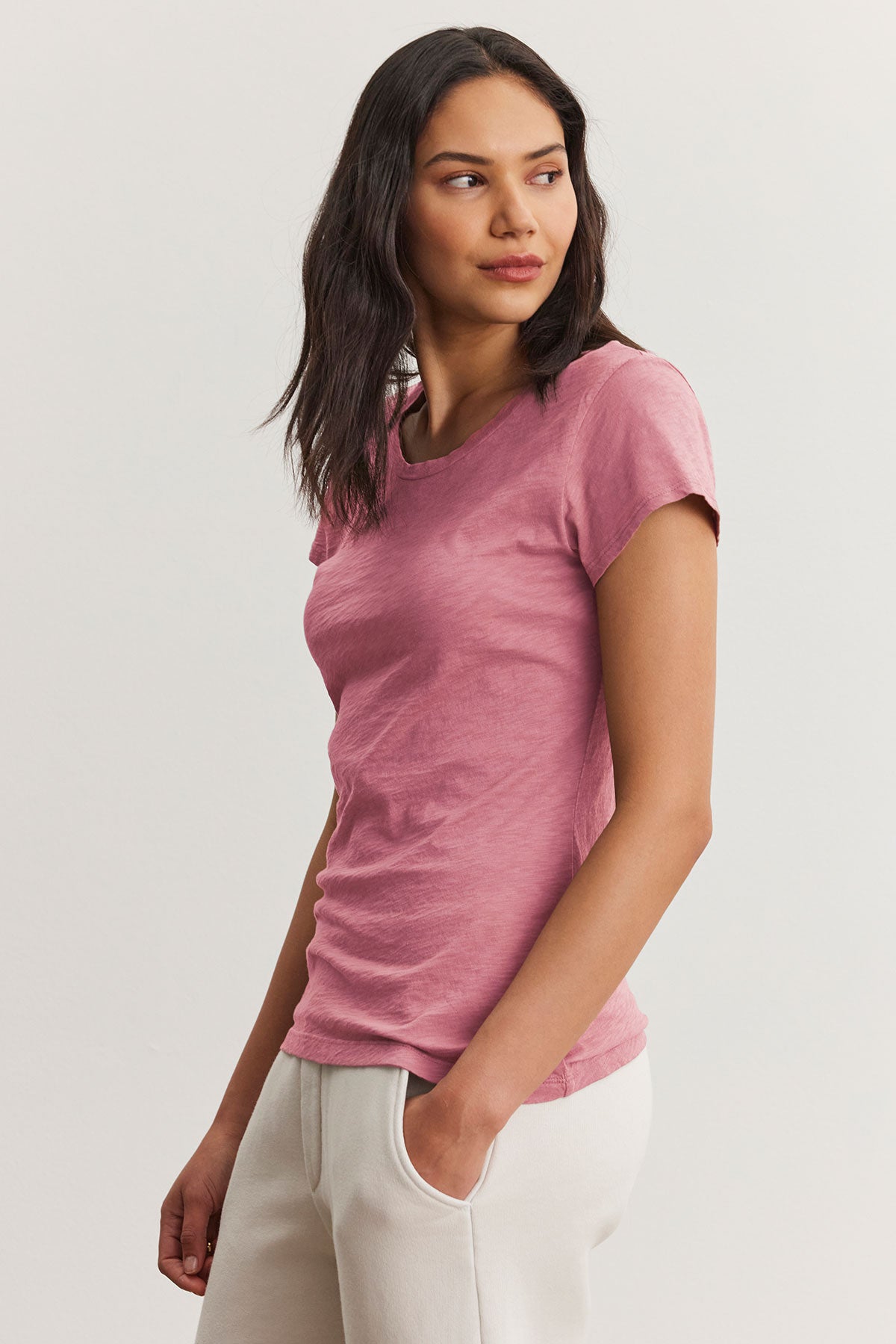   A person models the ODELIA TEE by Velvet by Graham & Spencer, a pink cotton slub fabric T-shirt with a classic crew neckline, paired with white pants against a plain backdrop. 