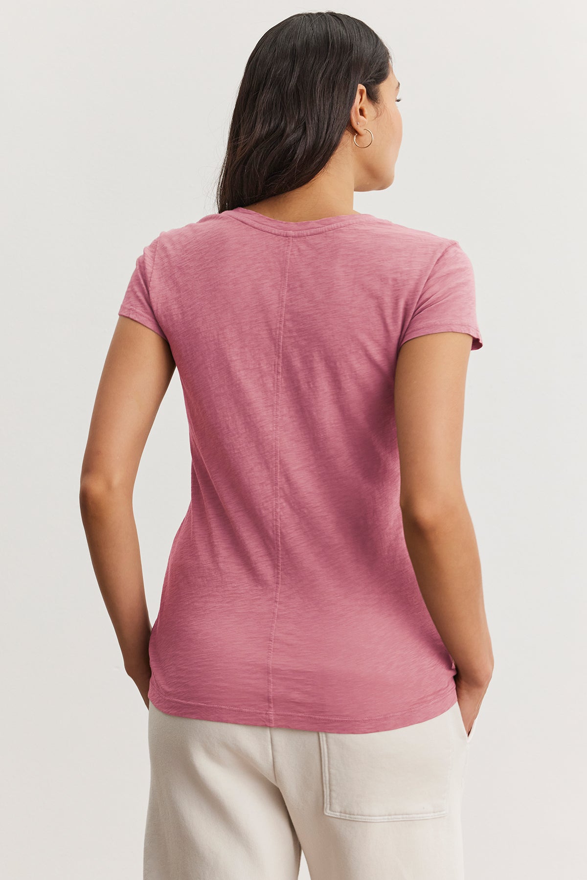 A person embodies the perfect wardrobe essential wearing an ODELIA TEE from Velvet by Graham & Spencer. It's a pink t-shirt featuring a classic crew neckline, paired with white pants, standing in a neutral posture.-38663157350593