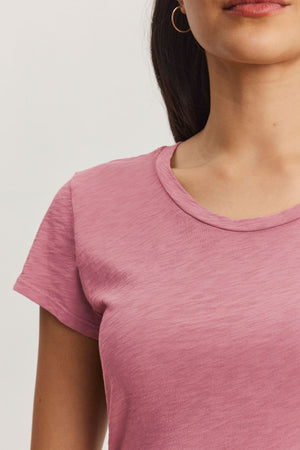 A person wears the ODELIA TEE by Velvet by Graham & Spencer, a pink short-sleeve shirt made from cotton slub fabric. The classic crew neckline of this wardrobe essential is highlighted as their upper body appears partially against a plain background, complete with a hoop earring.