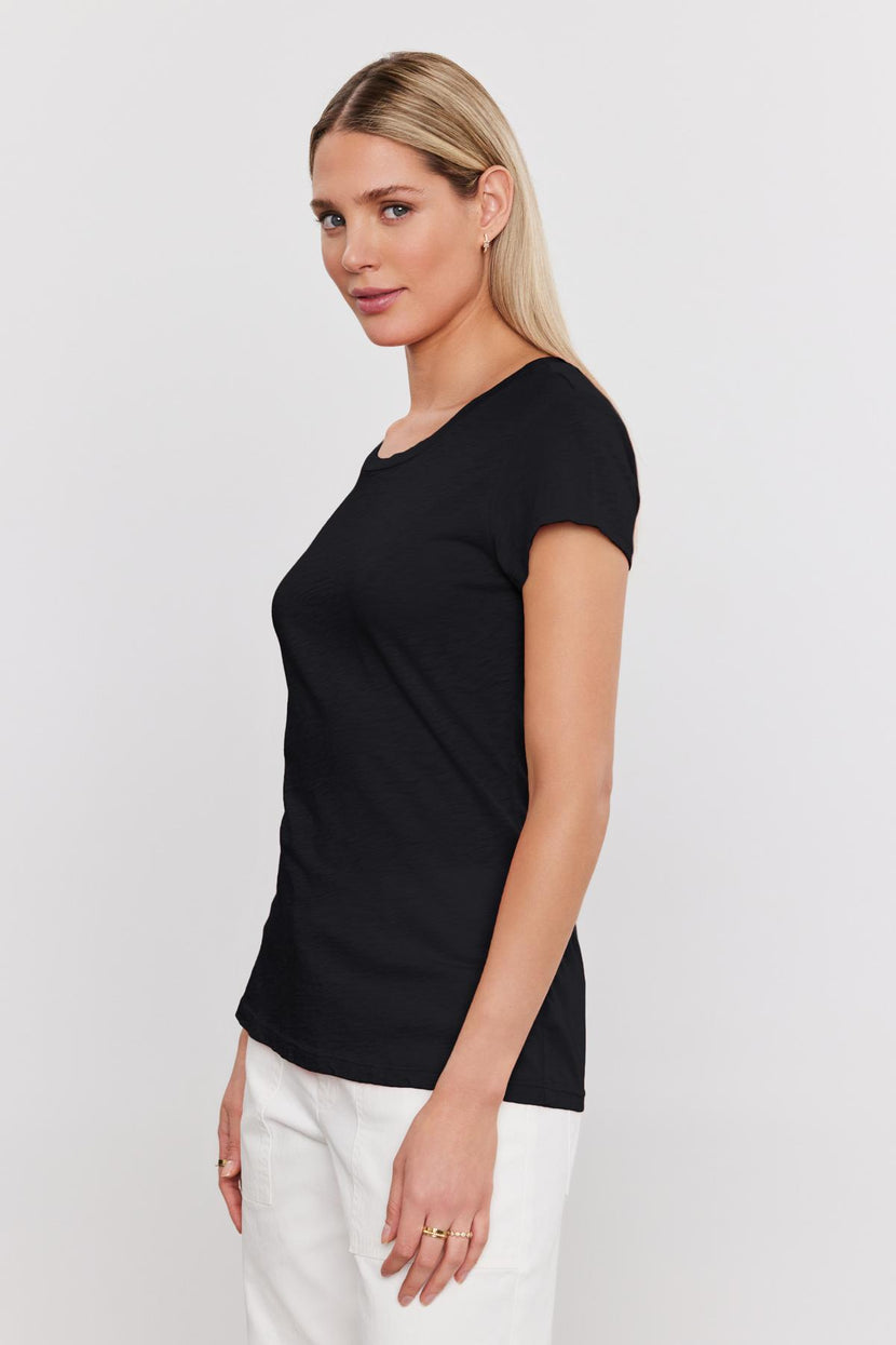 A woman with long blonde hair is wearing a fitted black t-shirt made from premium cotton slub and white pants, standing and looking to the side against a plain white background. Her ODELIA TEE by Velvet by Graham & Spencer adds a touch of timeless style to this wardrobe essential.