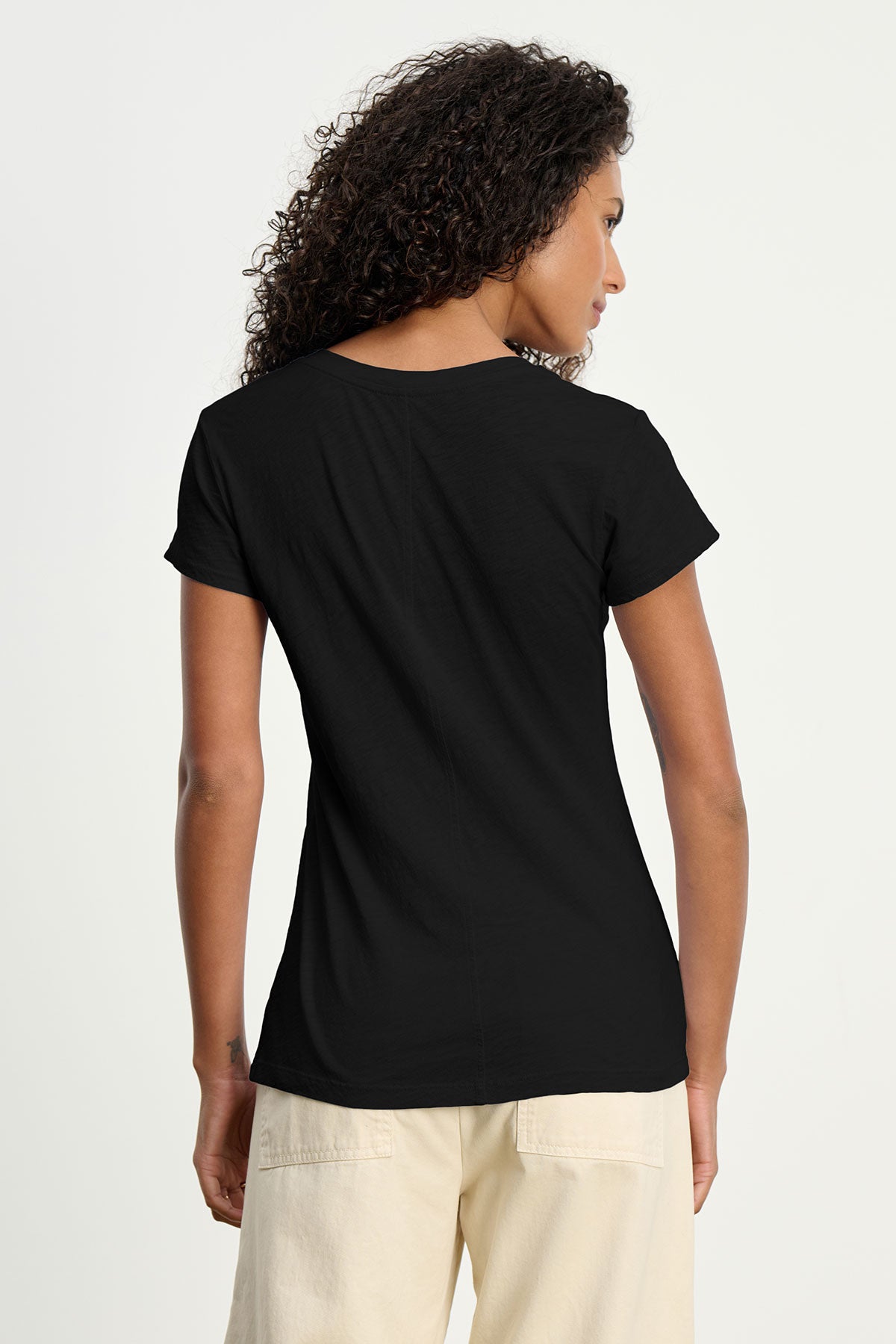   A person with curly hair is seen from the back, wearing the ODELIA TEE by Velvet by Graham & Spencer—a plain black premium cotton crew neck tee—paired with cream pants against a light background. 