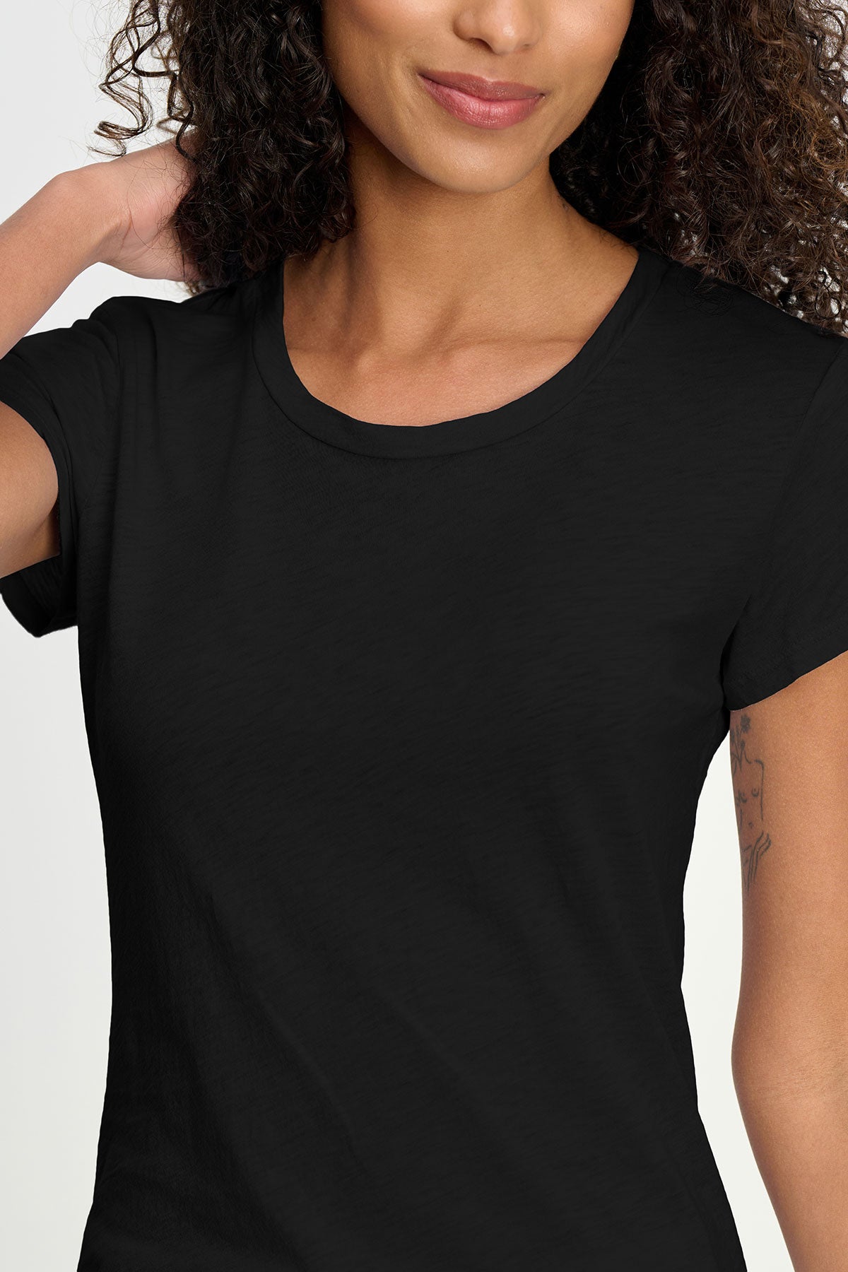   A woman with curly hair, partially smiling and touching her neck, wears the ODELIA TEE by Velvet by Graham & Spencer in black—a premium cotton essential with a classic crew neckline. 