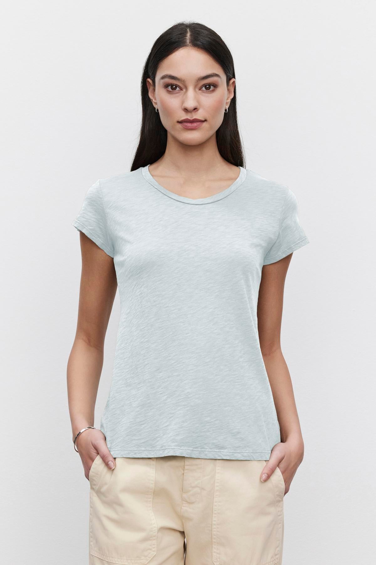   Young woman wearing a light blue Velvet by Graham & Spencer ODELIA COTTON SLUB CREW NECK TEE and beige pants, standing against a white background, looking directly at the camera. 