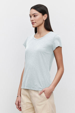 Woman standing against a white background, wearing a Velvet by Graham & Spencer ODELIA COTTON SLUB CREW NECK TEE in light blue and beige pants, looking to the side.