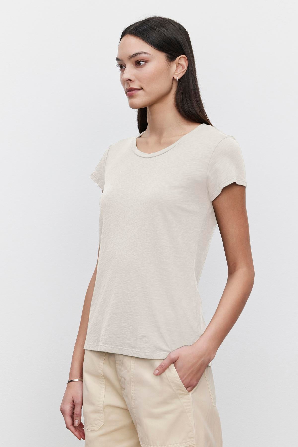   A woman with long dark hair is standing against a plain background, wearing a light beige ODELIA TEE made from premium cotton by Velvet by Graham & Spencer and light-colored pants, with her hand in her pocket. 