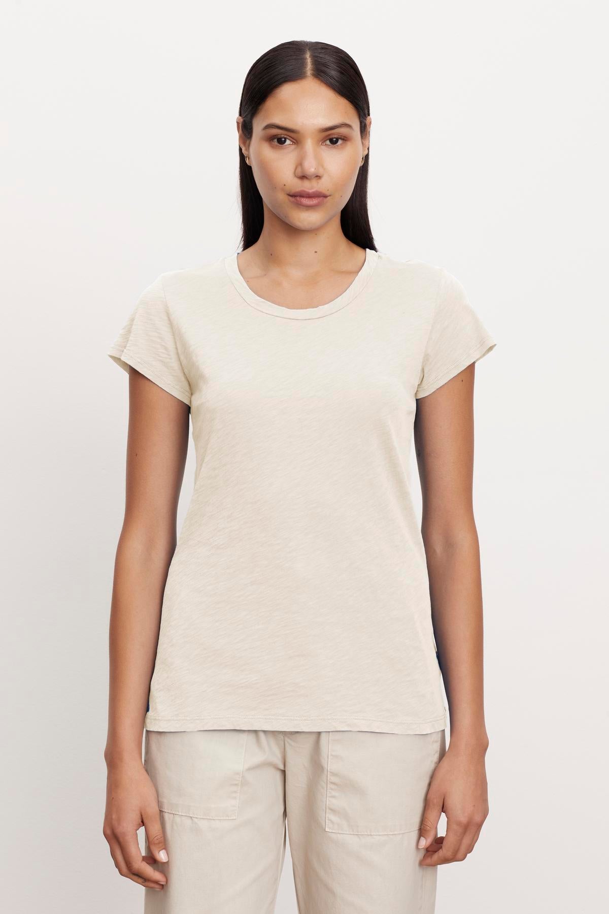 A person with long, straight hair stands facing the camera, wearing a short-sleeve, beige ODELIA TEE by Velvet by Graham & Spencer and light-colored pants against a plain background.-37249868431553