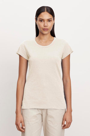 A person with long, straight hair stands facing the camera, wearing a short-sleeve, beige ODELIA TEE by Velvet by Graham & Spencer and light-colored pants against a plain background.
