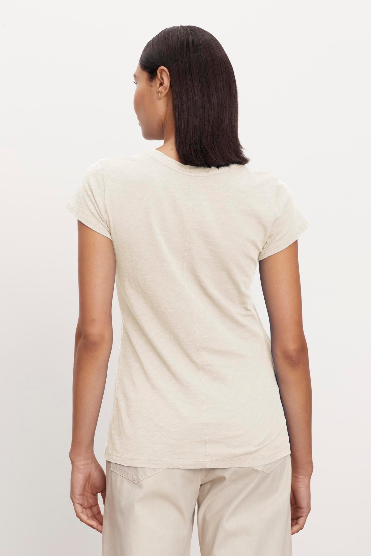 A person with straight, shoulder-length hair is facing away, wearing a light-colored Velvet by Graham & Spencer ODELIA TEE with a classic crew neckline and light-colored pants made from premium cotton against a plain background.-37249868497089