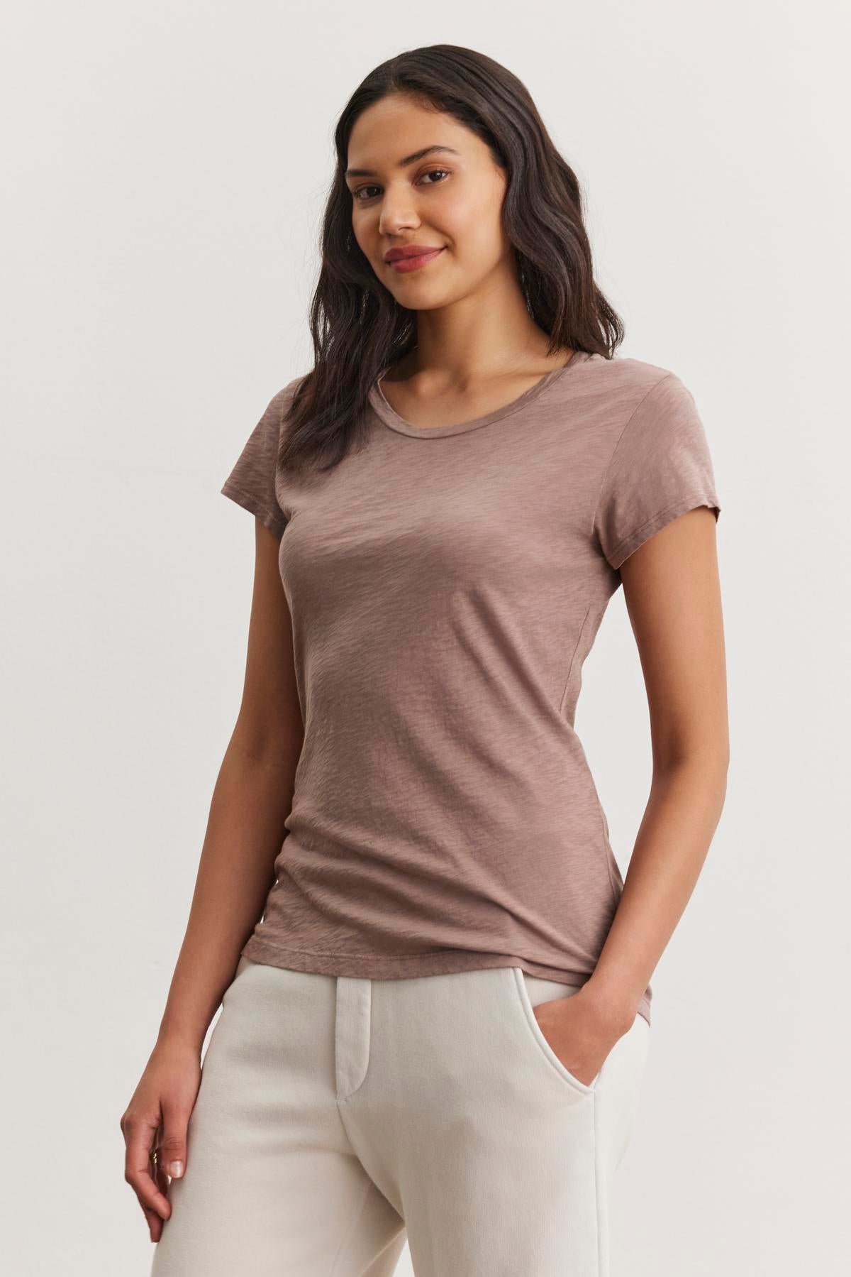   Amid a simple backdrop, a person stands with one hand in their pocket, wearing Velvet by Graham & Spencer's ODELIA TEE in brown slub cotton fabric and white pants. This wardrobe essential boasts a classic crew neckline ideal for any casual setting. 