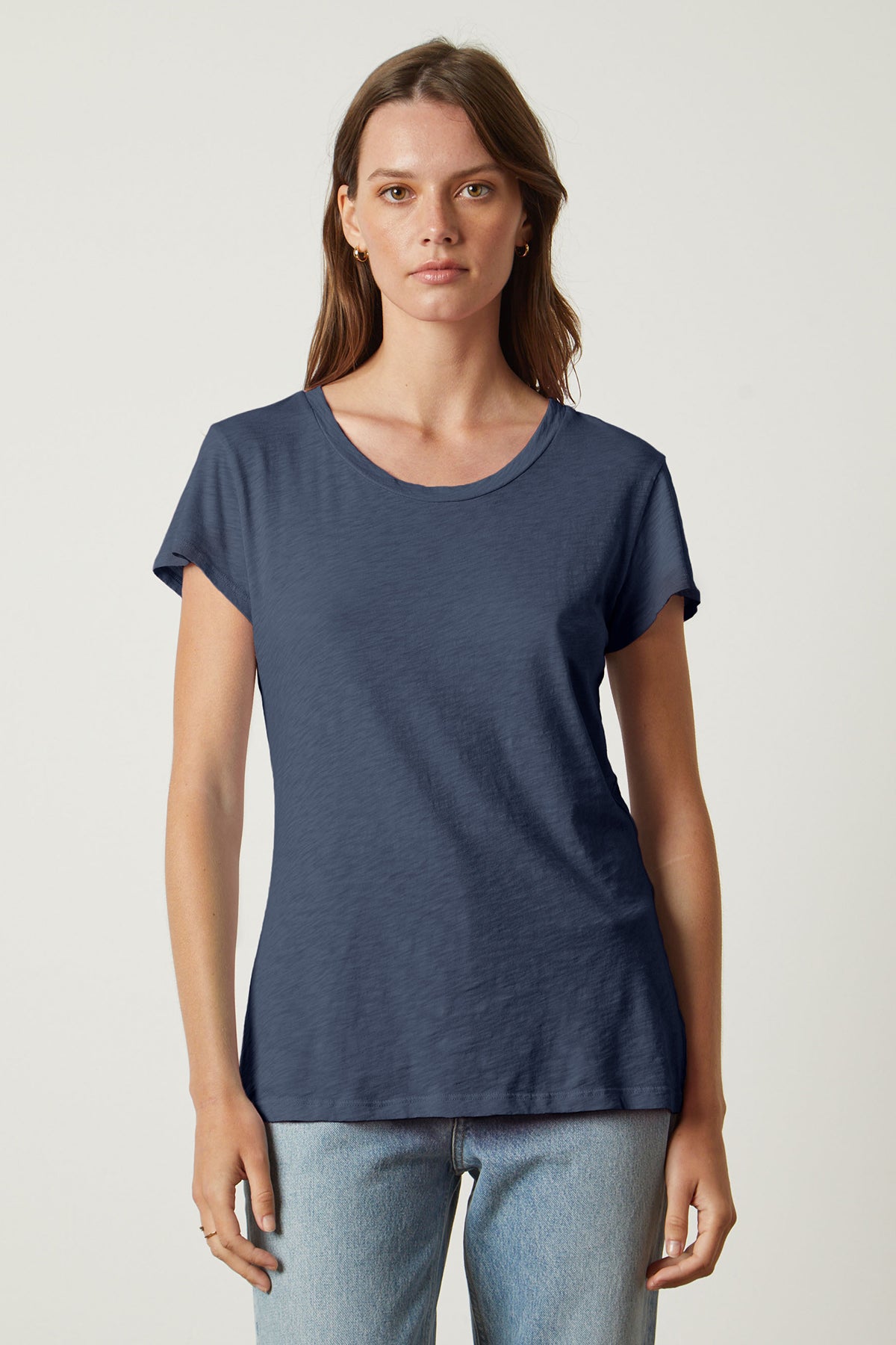   A woman wearing a plain, navy blue ODELIA TEE with a classic crew neckline by Velvet by Graham & Spencer stands against a light grey background. She has straight, shoulder-length brown hair and is wearing light blue jeans. This wardrobe essential is made from premium cotton, perfect for any casual occasion. 