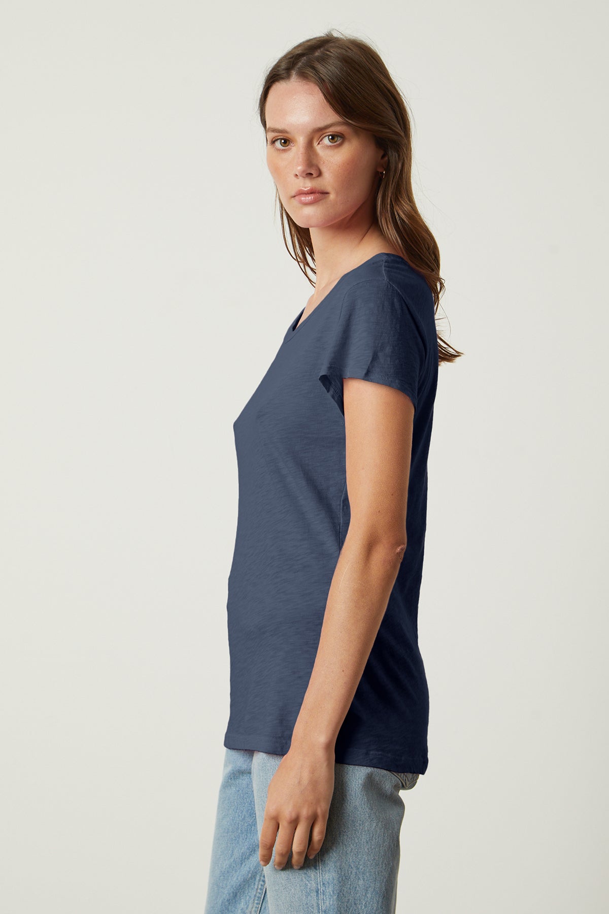 A woman with long brown hair is wearing a navy blue ODELIA TEE by Velvet by Graham & Spencer and light blue jeans, standing sideways and looking at the camera.-37250155446465
