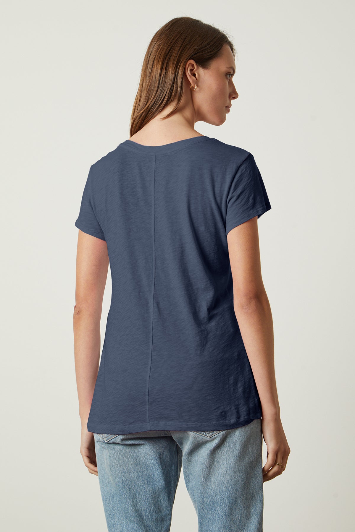 A person with long brown hair is standing and facing away from the camera, wearing a navy blue short-sleeve ODELIA TEE by Velvet by Graham & Spencer with a classic crew neckline and light blue jeans. This wardrobe essential showcases premium cotton for ultimate comfort.-37250155479233