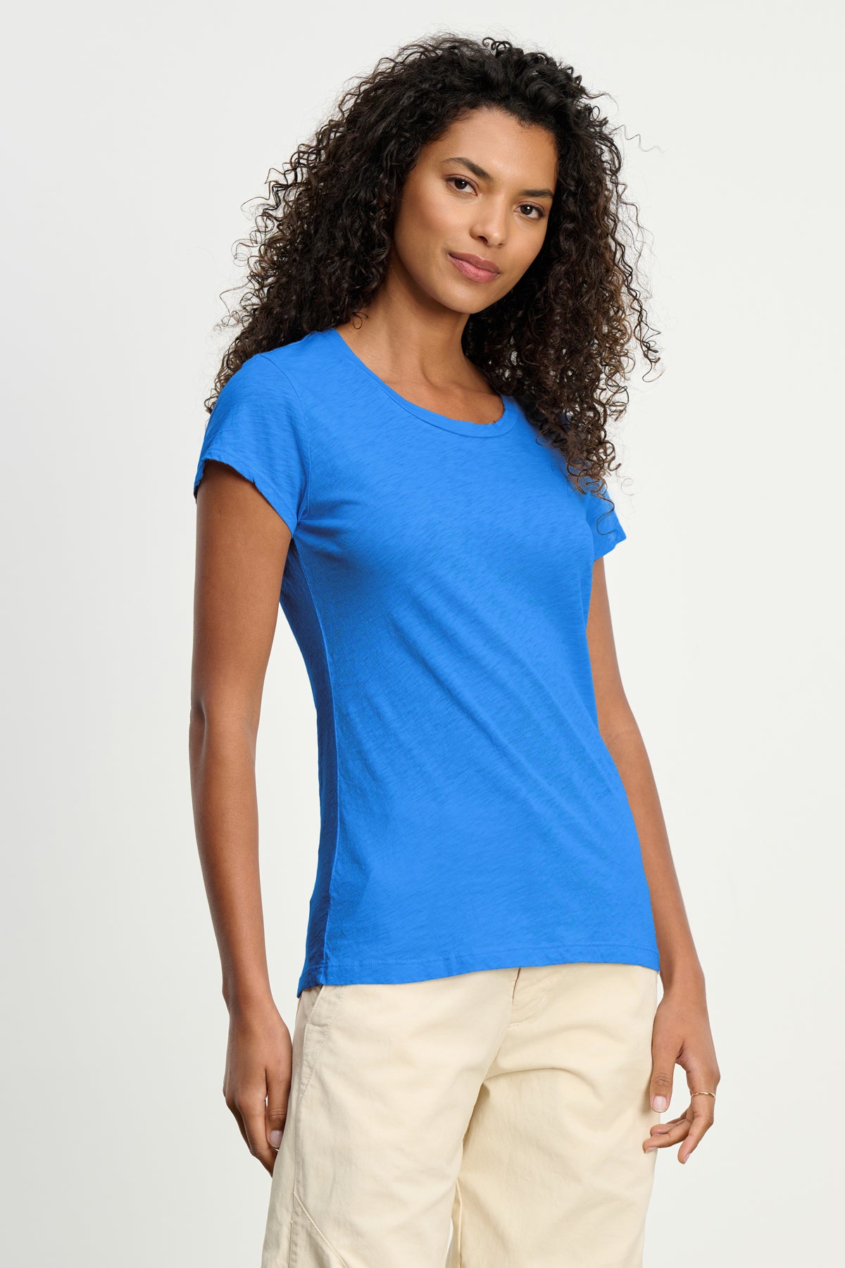 A woman with curly hair wears the Velvet by Graham & Spencer ODELIA TEE—a bright blue, short-sleeved crew neck T-shirt—with light-colored pants, standing against a plain light background.-39149216071873