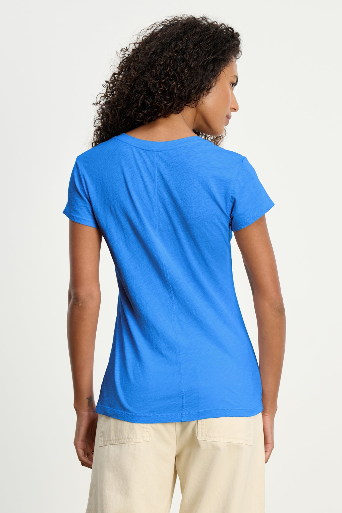 A woman with curly hair is seen from the back wearing the ODELIA TEE by Velvet by Graham & Spencer in blue soft cotton slub, paired with beige pants, against a plain background.-39149216104641