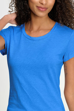A woman with curly hair smiles gently, wearing the ODELIA TEE by Velvet by Graham & Spencer—a bright blue, versatile tee crafted from soft cotton slub fabric for all-day comfort—standing against a neutral background.