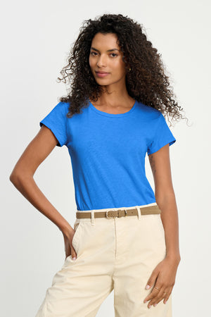 A woman with curly hair wears the ODELIA TEE by Velvet by Graham & Spencer in bright blue cotton slub, paired with beige pants, posing with one hand on her hip against a plain background.