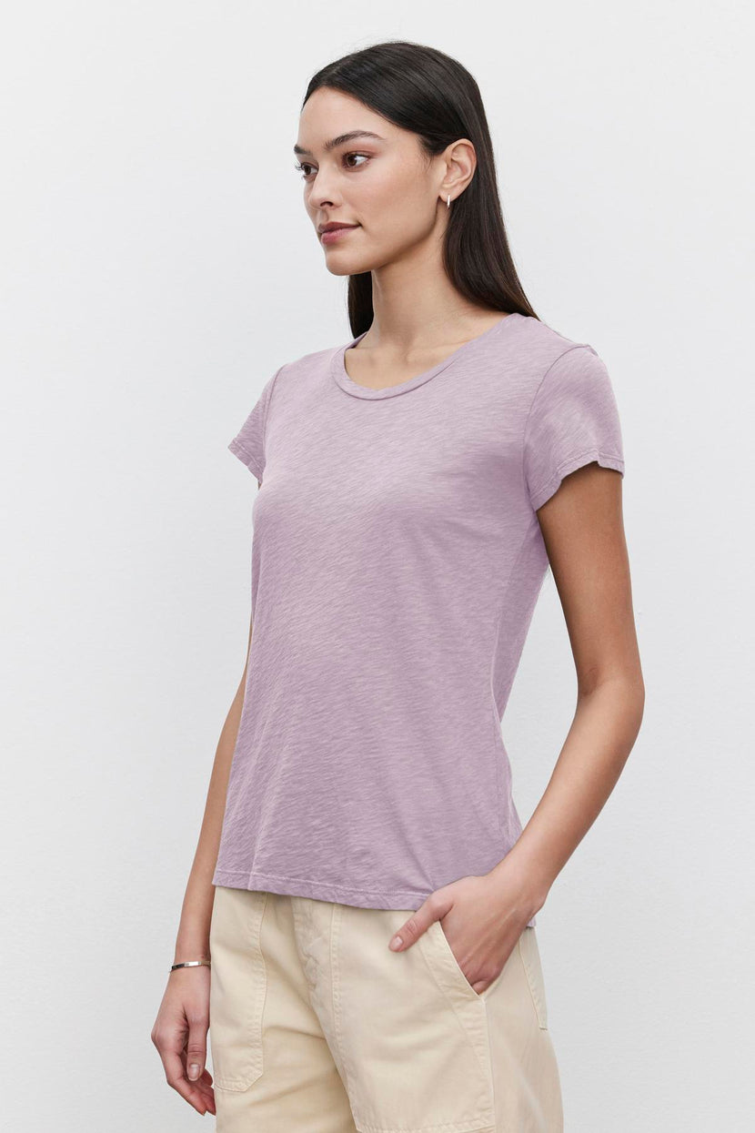 A person with long dark hair, wearing a short-sleeved lavender ODELIA TEE by Velvet by Graham & Spencer with a classic crew neckline and beige pants, stands against a plain white background.