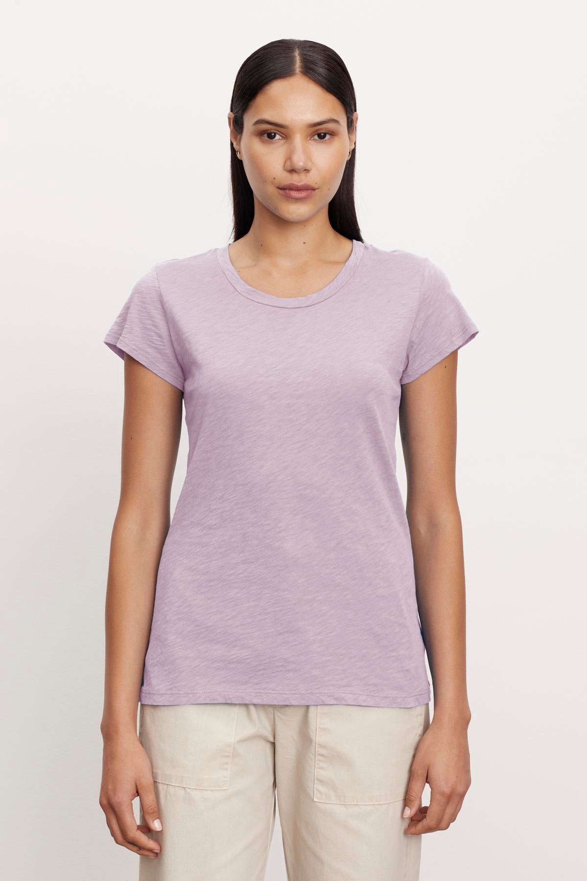 A woman with long, straight hair stands against a plain background wearing a short-sleeve, light purple ODELIA TEE by Velvet by Graham & Spencer with a classic crew neckline and light beige pants. Crafted from premium cotton, this wardrobe essential provides both comfort and style as she faces the camera with a neutral expression.-37249868529857
