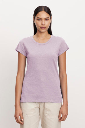 A woman with long, straight hair stands against a plain background wearing a short-sleeve, light purple ODELIA TEE by Velvet by Graham & Spencer with a classic crew neckline and light beige pants. Crafted from premium cotton, this wardrobe essential provides both comfort and style as she faces the camera with a neutral expression.