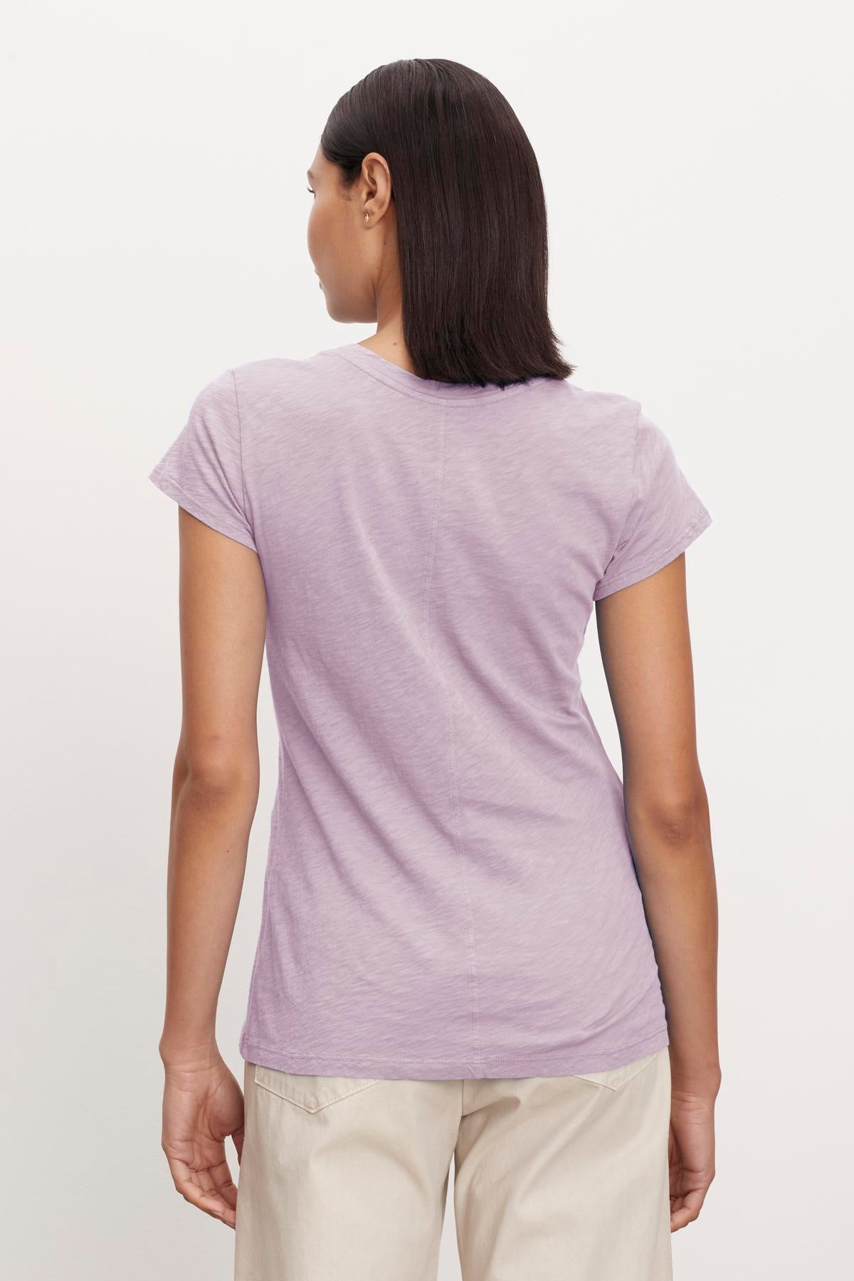   A person with short, dark hair stands facing away, wearing a light purple ODELIA TEE by Velvet by Graham & Spencer with a classic crew neckline and beige pants against a plain white background. The t-shirt is made from premium cotton, making it a wardrobe essential. 
