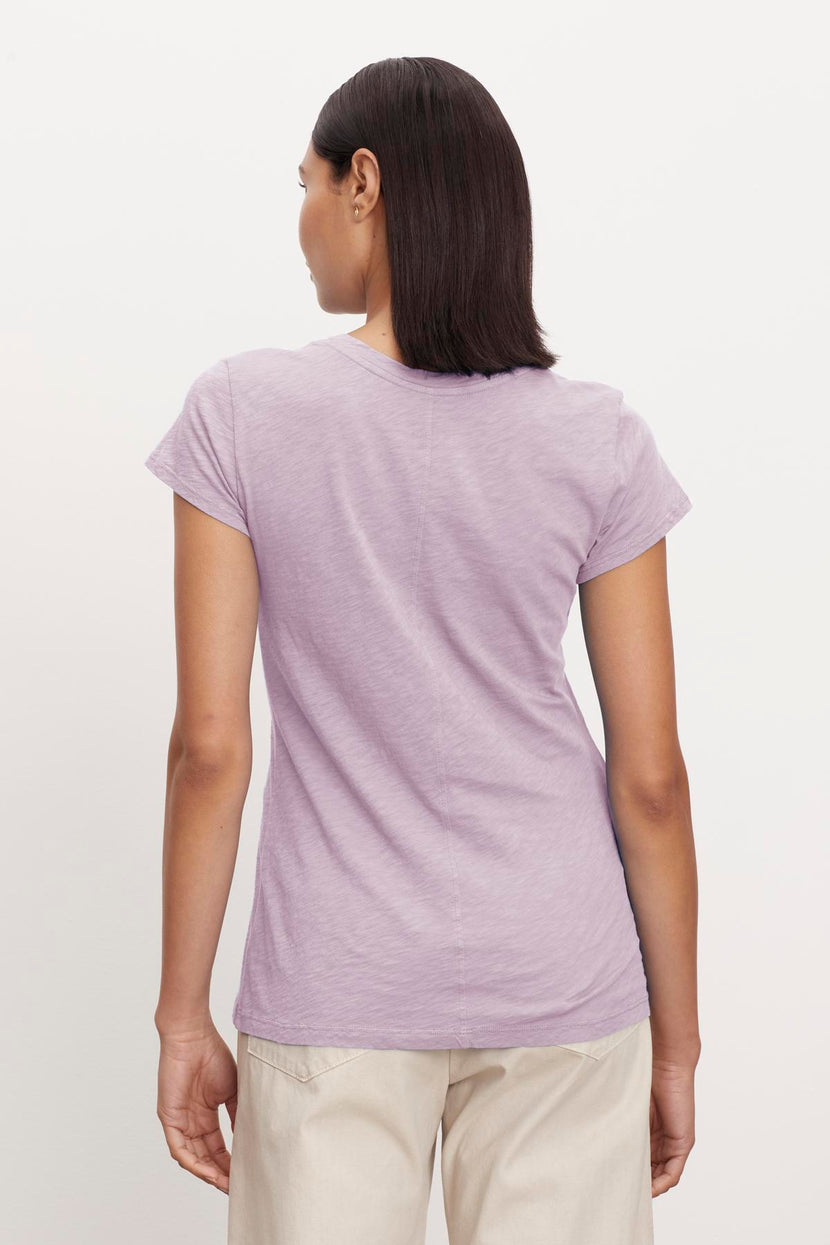 A person with short, dark hair stands facing away, wearing a light purple ODELIA TEE by Velvet by Graham & Spencer with a classic crew neckline and beige pants against a plain white background. The t-shirt is made from premium cotton, making it a wardrobe essential.