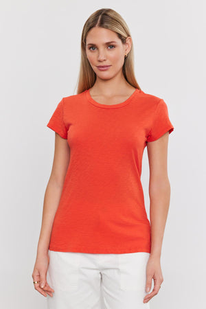 A woman standing against a white background, wearing a bright orange ODELIA COTTON SLUB CREW NECK TEE from Velvet by Graham & Spencer and white pants, looking directly at the camera.