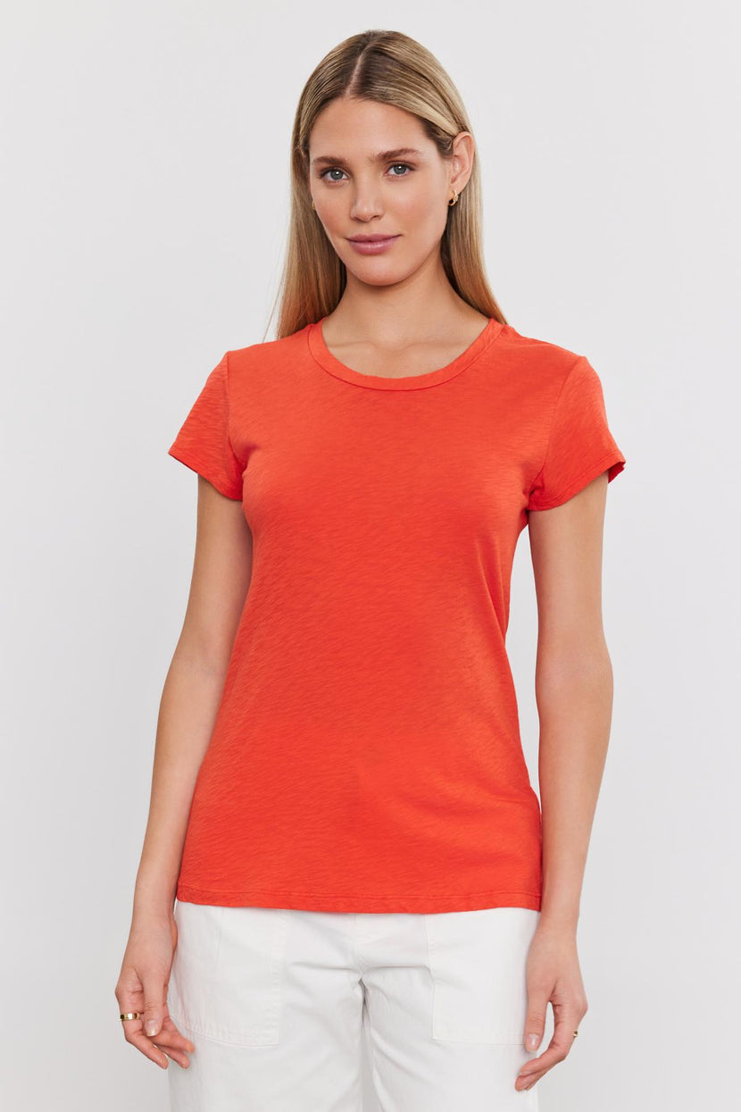 A woman with long blonde hair wearing an orange ODELIA TEE by Velvet by Graham & Spencer with a classic crew neckline and white pants stands against a plain white background.