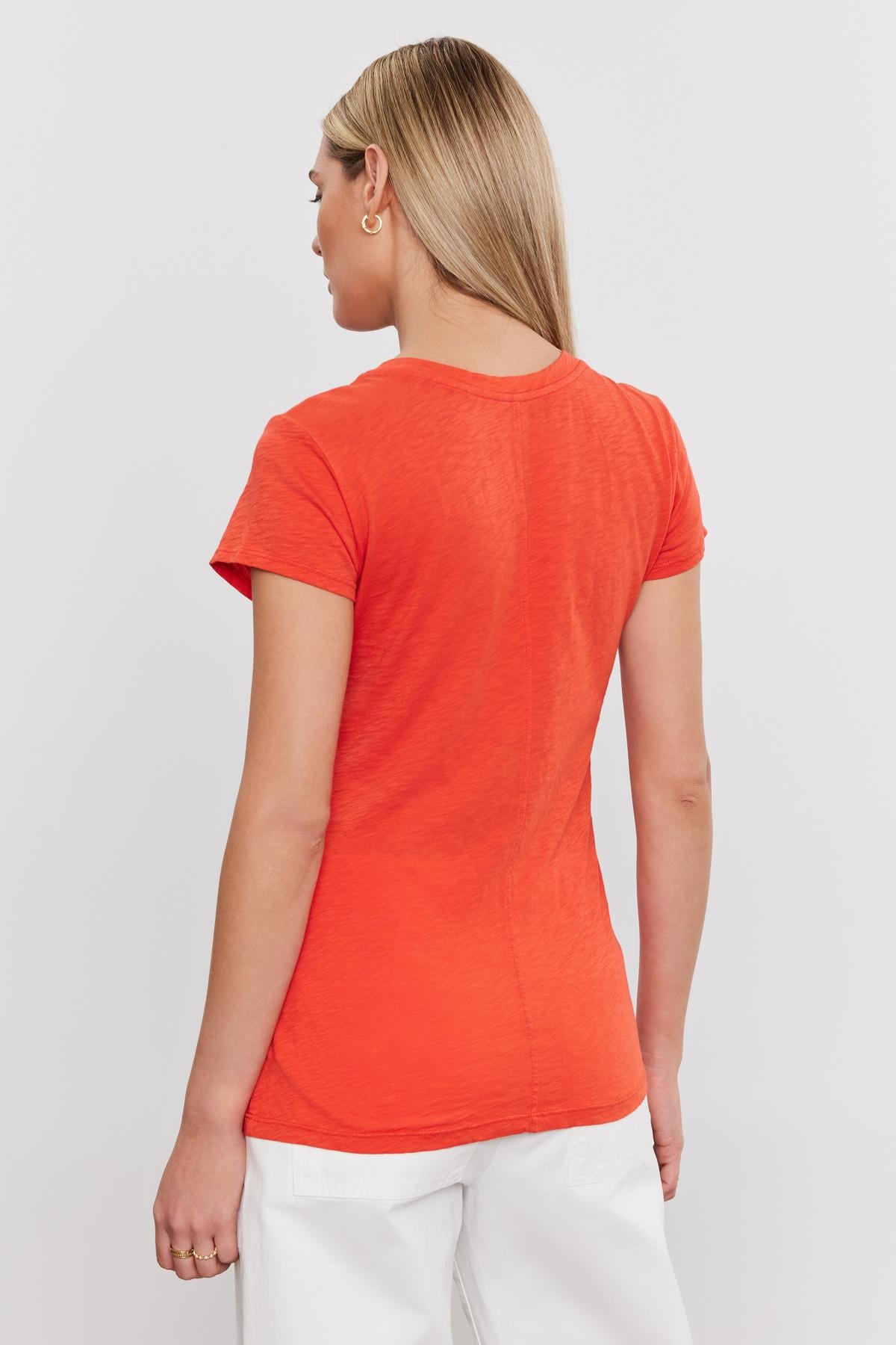   Woman in a bright orange ODELIA COTTON SLUB CREW NECK TEE by Velvet by Graham & Spencer and white pants, viewed from the back. Her hair is styled in a ponytail. 