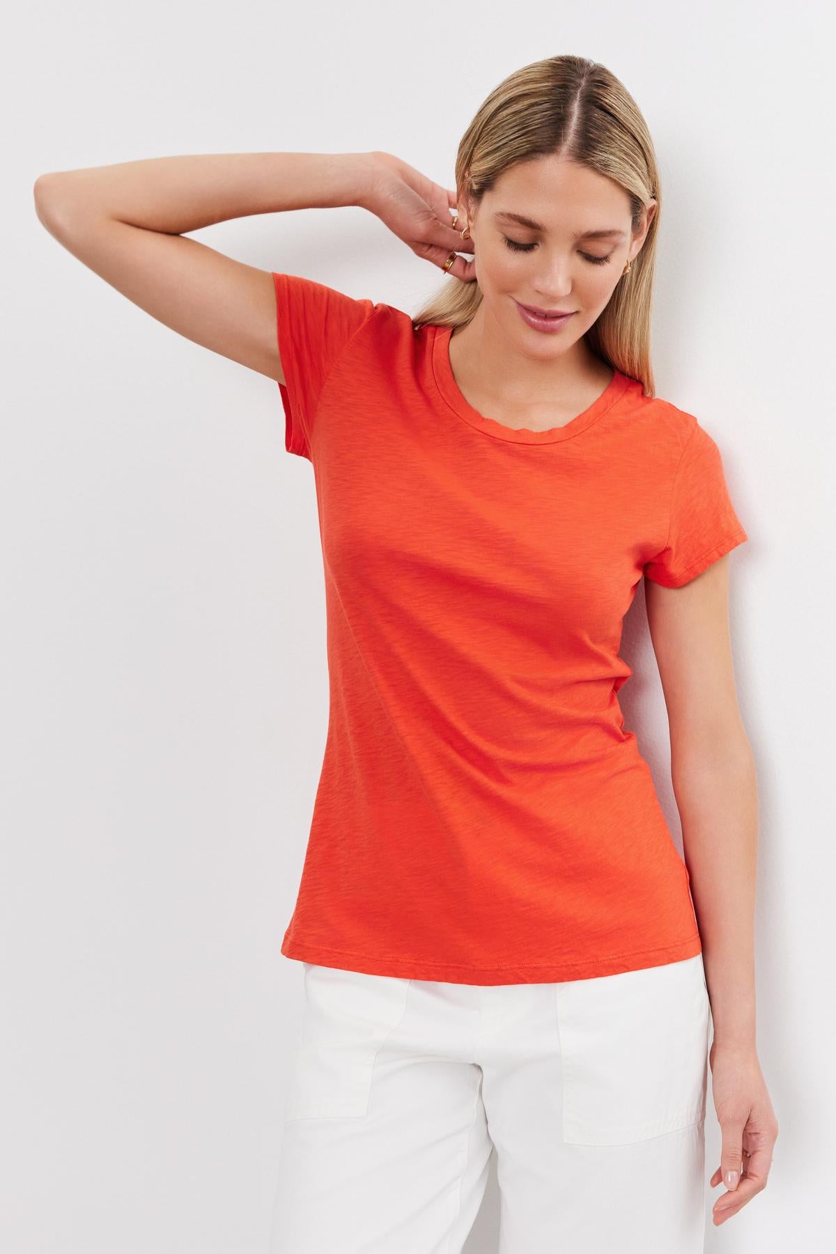   A young woman wearing a bright orange Velvet by Graham & Spencer ODELIA COTTON SLUB CREW NECK TEE and white pants, posing with her hand on her neck against a white background. 