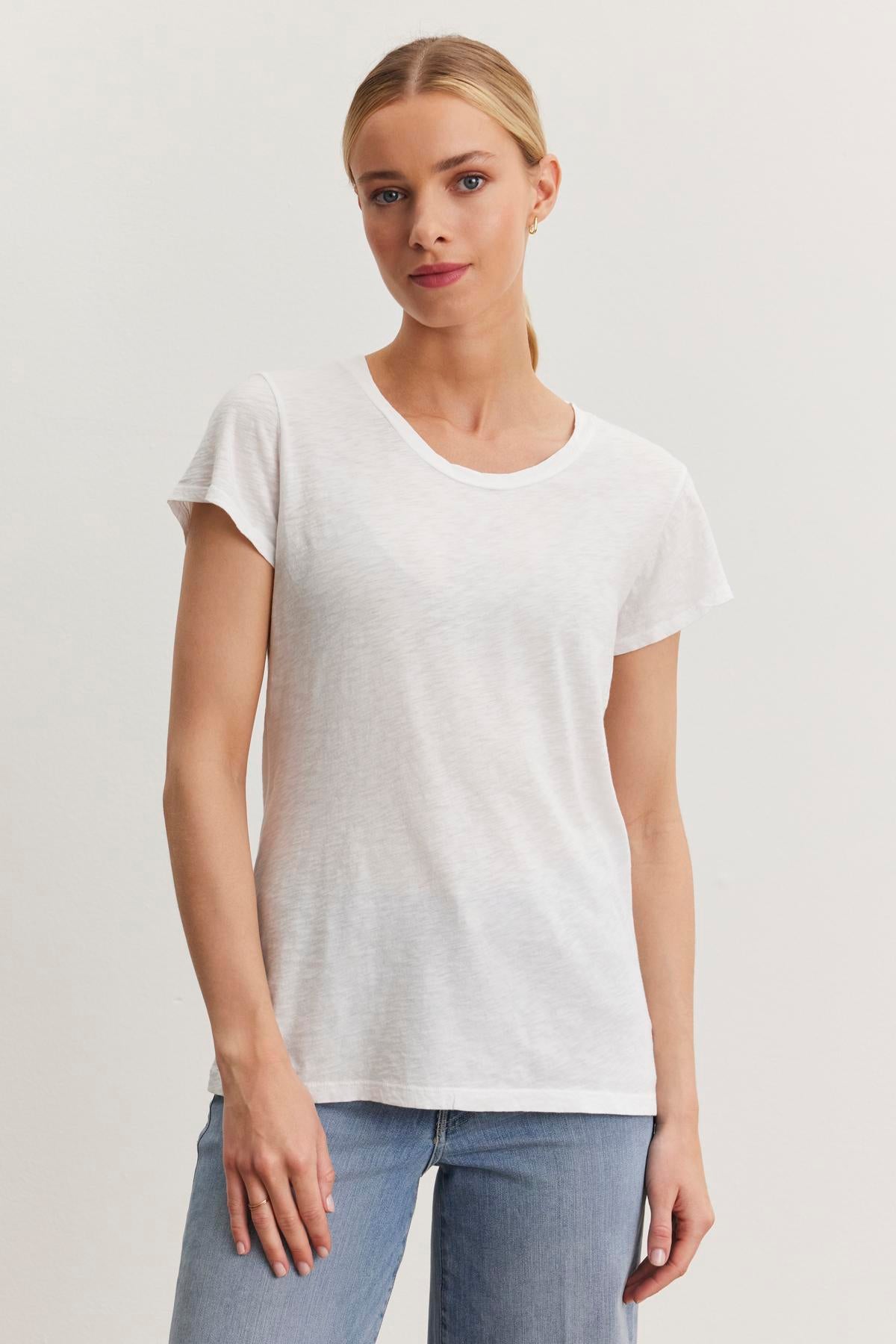 A woman with blonde hair stands against a plain background, wearing the ODELIA TEE by Velvet by Graham & Spencer, a versatile white short-sleeve t-shirt made from premium cotton slub fabric, and blue jeans.-37366925918401