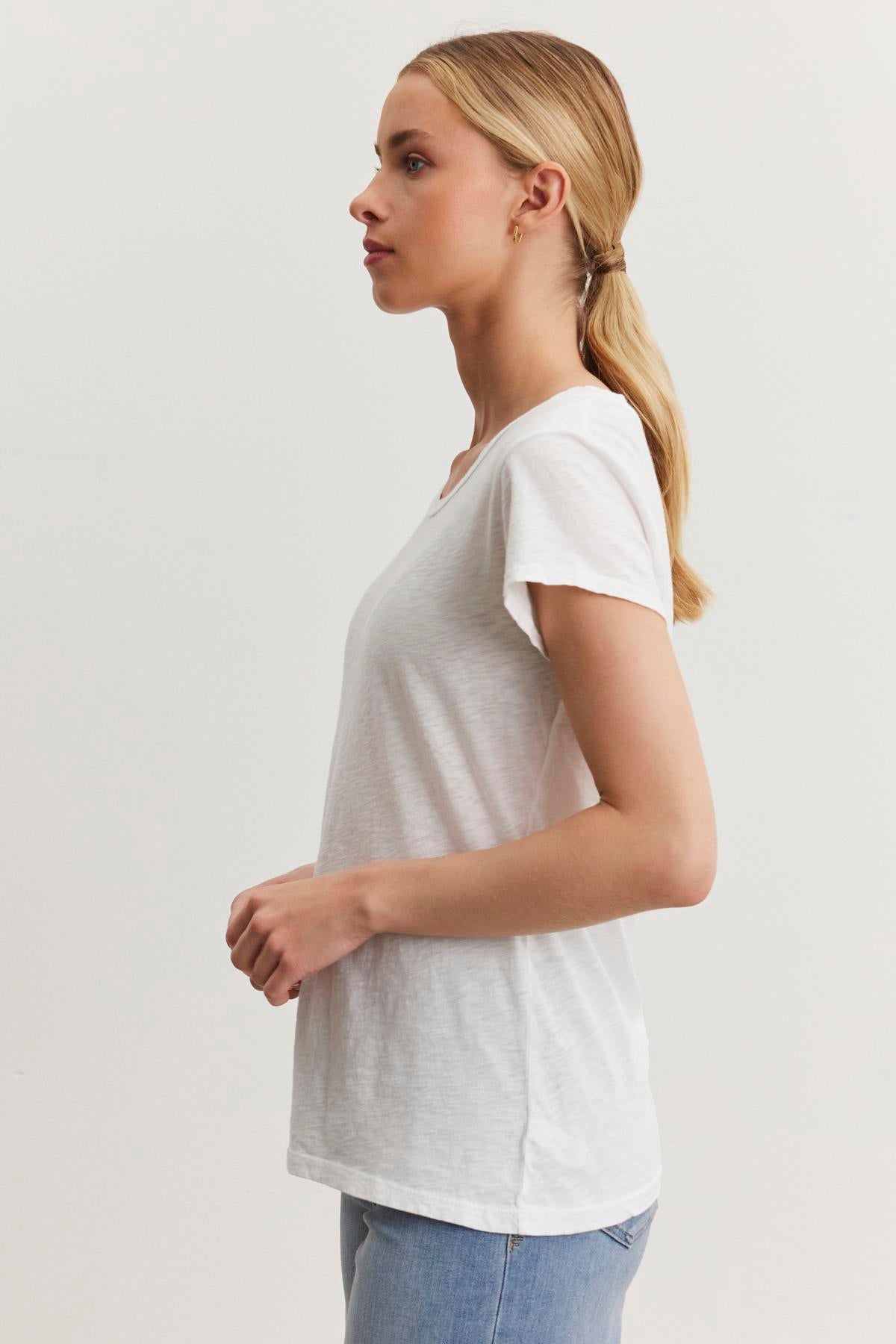   A woman with long blonde hair tied in a low ponytail is standing in profile view, wearing the ODELIA TEE from Velvet by Graham & Spencer made from premium cotton slub fabric and light blue jeans. This versatile wardrobe essential perfectly complements the plain white background as she faces left. 