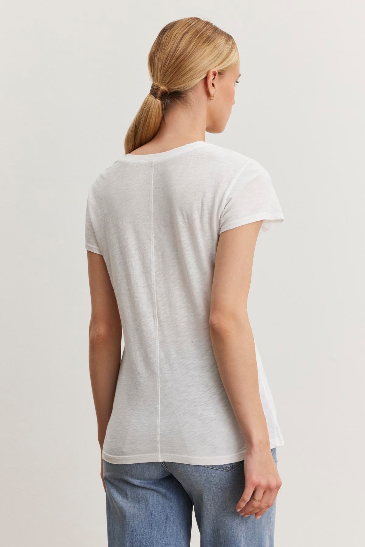   A person with blonde hair in a ponytail is facing away, wearing the Velvet by Graham & Spencer ODELIA TEE made from premium cotton slub fabric and blue jeans. 