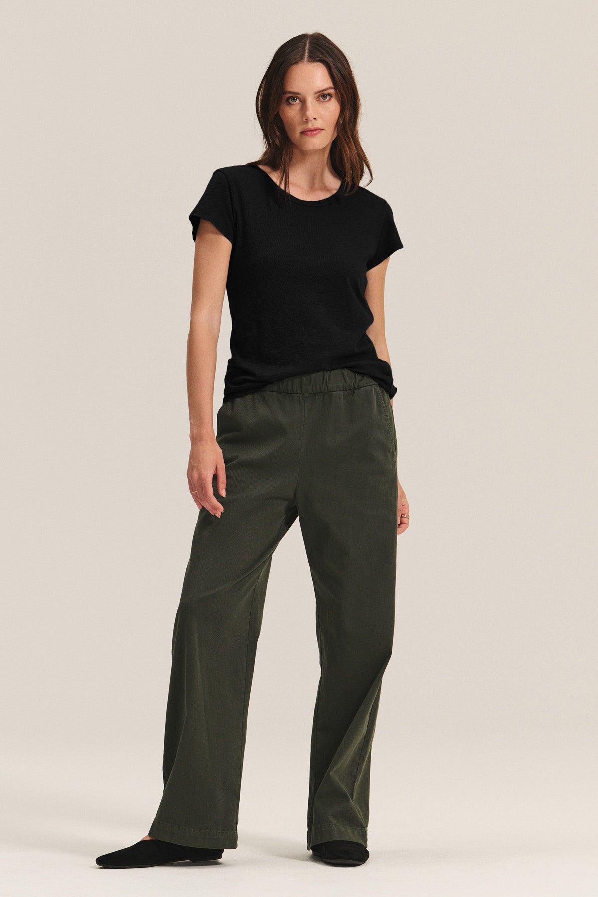 A woman stands against a plain background wearing the Velvet by Graham & Spencer ODELIA TEE in black, paired with olive green loose-fitting pants and black shoes.-40292940742849